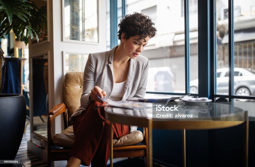 Woman reading a magazine at cafe Young woman reading a magazine while sitting at restaurant table. Caucasian female in casuals relaxing at a cafe. Magazine - Publication Stock Photo