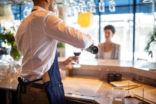 Bartender serving a drink to businesswoman in bar Bartender pouring wine into a glass with a woman sitting at bar counter. Barman serving a drink to businesswoman in bar. cocktail bar busy stock pictures, royalty-free photos & images