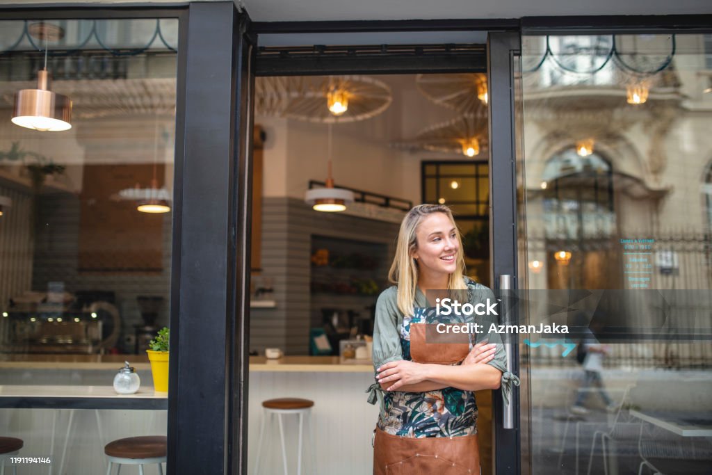 Young Cafe Owner Standing with Arms Crossed at Front Door Hopeful Argentine female cafe owner standing in apron with arms crossed at open front door and looking away from camera. Store Stock Photo