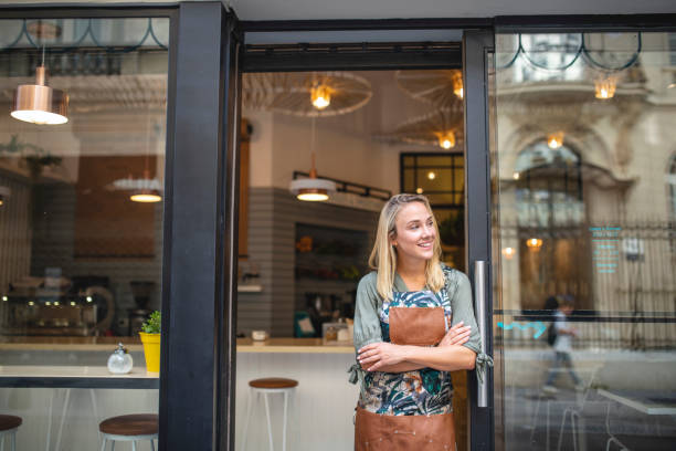 Young Cafe Owner Standing with Arms Crossed at Front Door Hopeful Argentine female cafe owner standing in apron with arms crossed at open front door and looking away from camera. retail place stock pictures, royalty-free photos & images