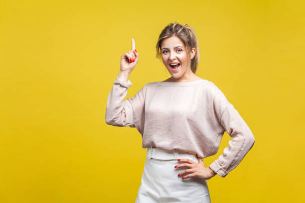 portrait of inspired beautiful young woman with fair hair in casual beige blouse, isolated on yellow background - finger raised imagens e fotografias de stock