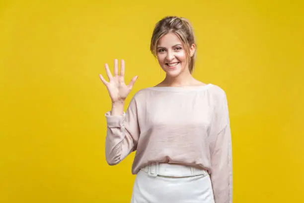 Hi there! Portrait of friendly young beautiful woman with fair hair in casual blouse standing looking at camera with raised arm, gesturing welcome. indoor studio shot isolated on yellow background