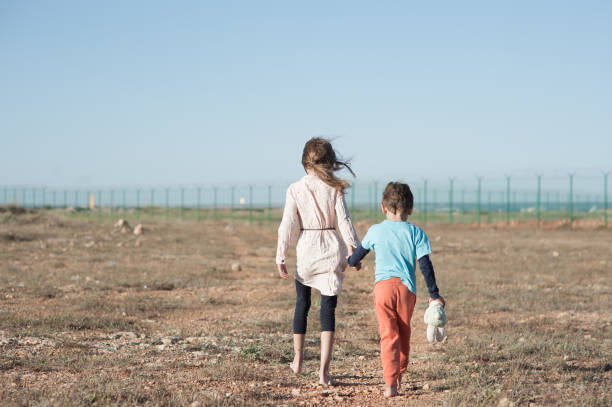 two poor children family brother with toy and thin sister refugee illegal immigrant walking barefooted through hot desert towards state border with barbed fence wire two poor children family brother and sister refugee illegal immigrant walking barefooted through hot desert towards state border with barbed fence wire refugee camp stock pictures, royalty-free photos & images