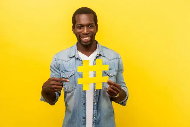Photo of Viral hashtag and successful blogging. Portrait of happy man pointing at large yellow hash symbol. indoor studio shot isolated, yellow background