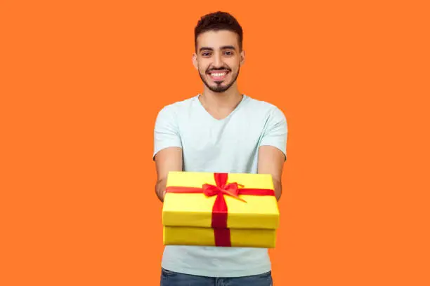 Photo of Portrait of happy generous brunette man giving gift box to camera. indoor studio shot isolated on orange background