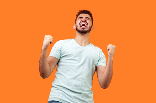Bottom view of ecstatic motivated brunette man with beard in white t-shirt standing with raised fists and shouting for joy, winner excited for success. indoor studio shot isolated on orange background