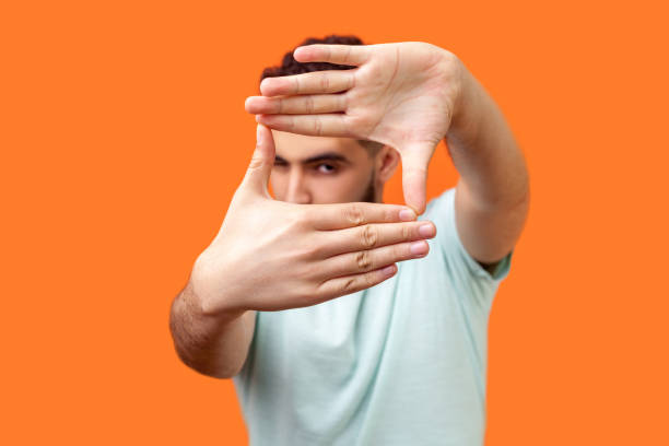 Portrait of attentive curious brunette man focusing through photo frame made of fingers. indoor studio shot isolated on orange background Portrait of attentive curious brunette man in casual white t-shirt looking at camera with one eye, focusing through photo frame made of fingers. indoor studio shot isolated on orange background viewpoint photos stock pictures, royalty-free photos & images