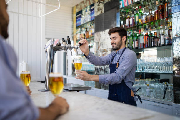 Bartender filing beer in a glass from tap Bartender pouring beer in a glass. Barman filling a beer in a glass from the tap with customer sitting at the counter. serving food and drinks stock pictures, royalty-free photos & images