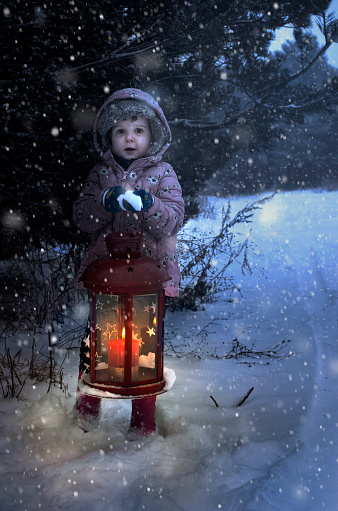 Child hold christmas lamp with glass and candle inside in the night to pine tree. Snowing around the lamp. Snowflakes flying in the night.