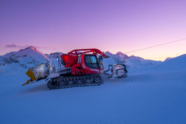 young man standing in front of a snow groomer in the mountains at evening