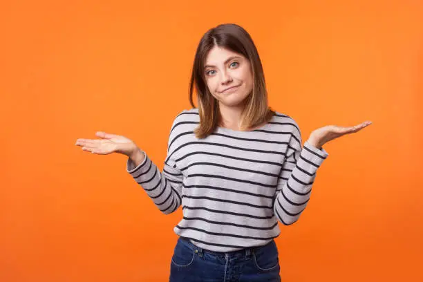 Don't know! Portrait of uncertain confused brunette woman in long sleeve shirt standing having doubts while raised hands, clueless facial expression. indoor studio shot isolated on orange background