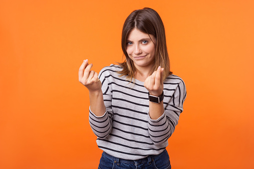 Portrait of smart beautiful woman with brown hair in long sleeve striped shirt standing showing money gesture with fingers, looking cunning at camera. indoor studio shot isolated on orange background