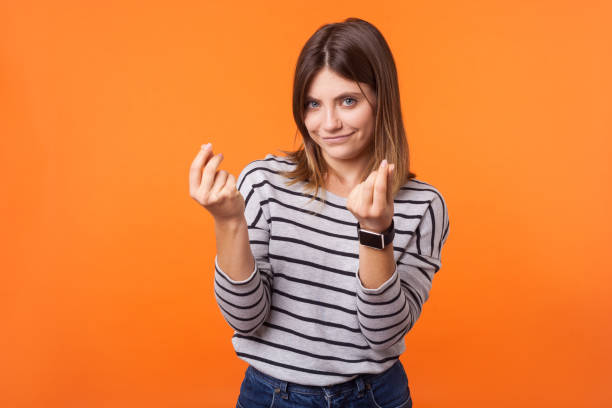 Portrait of smart beautiful woman with brown hair in long sleeve striped shirt. indoor studio shot isolated on orange background Portrait of smart beautiful woman with brown hair in long sleeve striped shirt standing showing money gesture with fingers, looking cunning at camera. indoor studio shot isolated on orange background italian ethnicity stock pictures, royalty-free photos & images