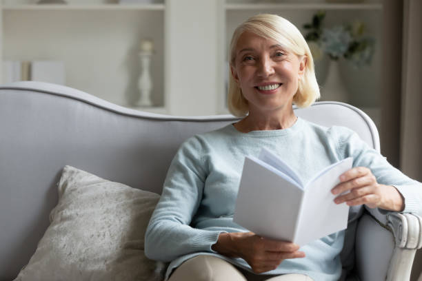 istock Aged woman sitting on couch holding book looking at camera 1191216457