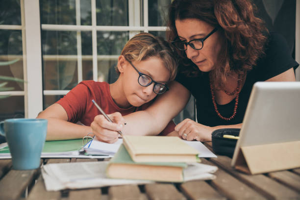 giovane studente che fa i compiti a casa con libri di scuola, giornale e pad digitale aiutato da sua madre. mamma che scrive sul copilota che insegna a suo figlio. istruzione, stile di vita familiare, concetto di homeschooling - home schooling foto e immagini stock