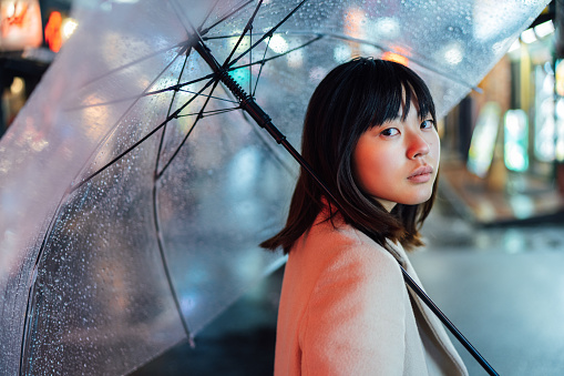 A portrait of a young woman on a rainy day at night in the street while holding an umbrella and looking at camera,.