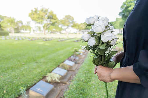 Mourning young woman holding white flowers at her family grave in beautiful green cemetery.