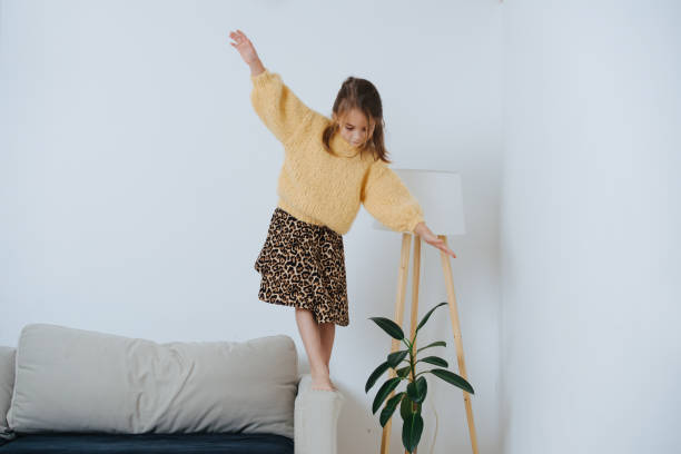 Little girl in sweater walking on a armrest of a couch, keeping balance. At home Brunette little girl in yellow knitted sweater and leopard skirt walking on an armrest of a couch at home. Her arms outstretched as she tries to maintain balance. couch arm rest stock pictures, royalty-free photos & images