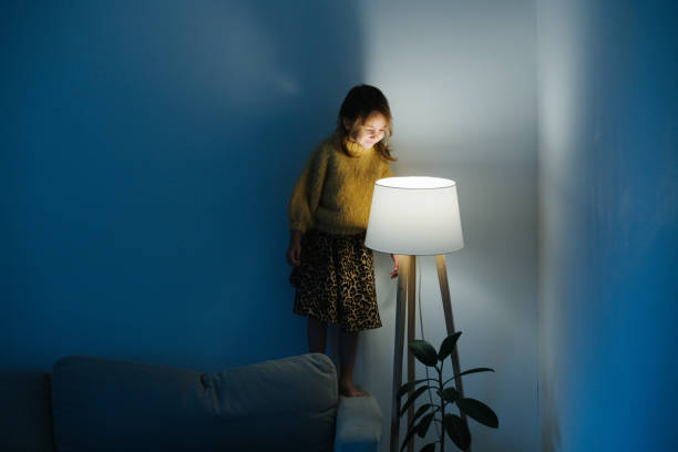Little girl in sweater standing on a couch looking at nigth light at home. Brunette little girl in yellow knitted sweater and leopard skirt standing on a backseat of a couch at home. She's looking at dim night light from above. Her face is lit. turning on or off photos stock pictures, royalty-free photos & images