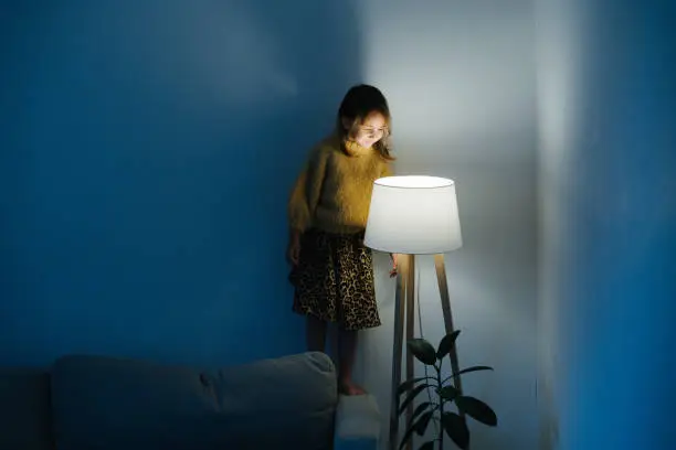 Photo of Little girl in sweater standing on a couch looking at nigth light at home.