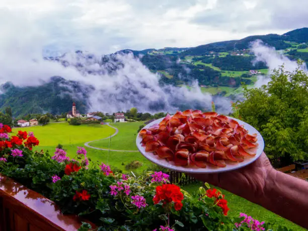 Hand holding plate of speck with amazing alpine landscape in the background. In Sant'Osvaldlo, Castelrotto, Dolomites, northern Italy