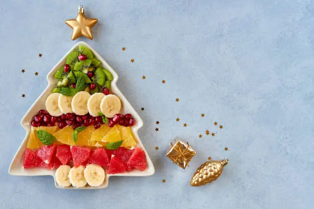 Photo of Christmas background with fruit salad in fir tree shaped plate and holiday decoration. Top view flat lay