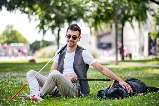 Young blind man with white cane and guide dog sitting in park in city, resting.