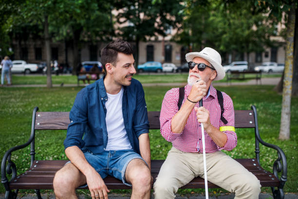 Young man and blind senior with white cane sitting on bench in park in city. A young man and blind senior with white cane sitting on bench in park in city, talking. aids walk stock pictures, royalty-free photos & images