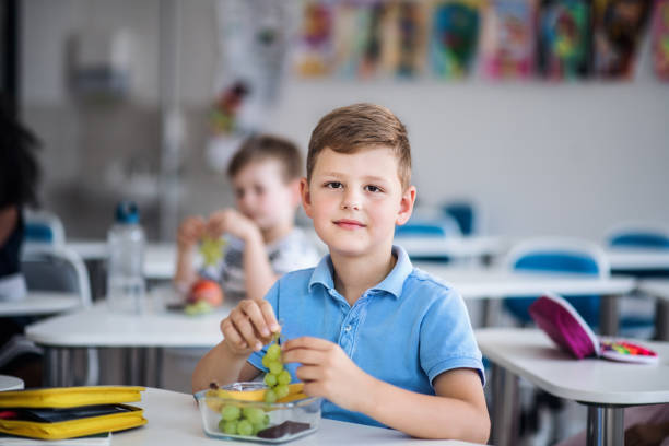 A small school boy sitting at the desk in classroom, eating grapes. A small happy school boy sitting at the desk in classroom, eating grapes fruit. food elementary student healthy eating schoolboy stock pictures, royalty-free photos & images