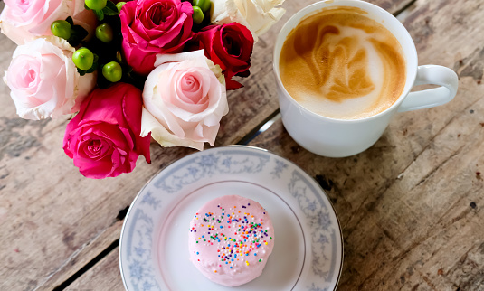 White cup of coffee latte, vanilla cake and pink roses on wooden table. Lifestyle concept.