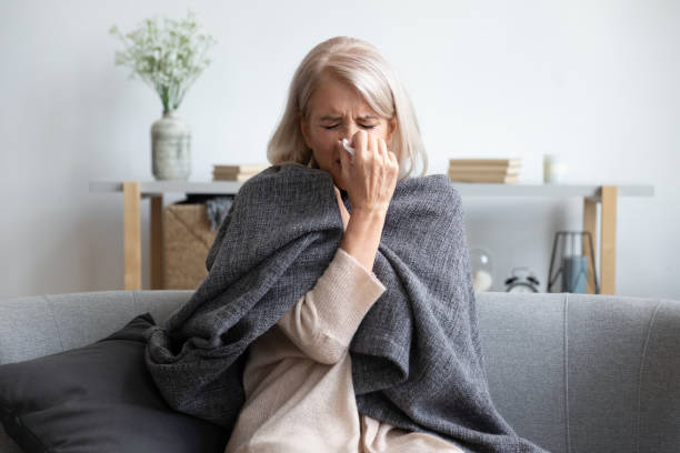 Aged sick woman sneezing holding napkin blow out runny nose Middle-aged 50s sick frozen woman seated on sofa in living room covered with warm plaid sneezing holding paper napkin blow out runny nose feels unhealthy, seasonal cold, weakened immune system concept influenza stock pictures, royalty-free photos & images