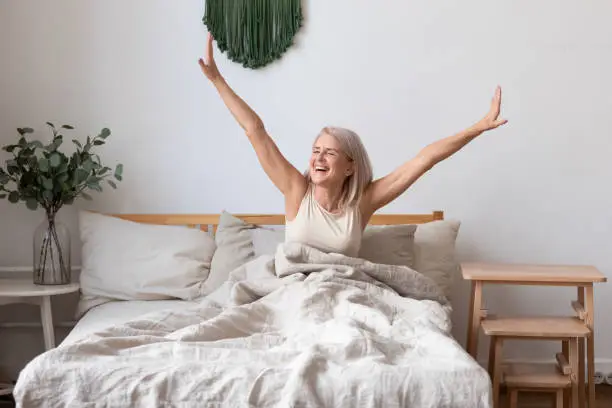 Photo of Aged female waking up in morning stretches seated in bed