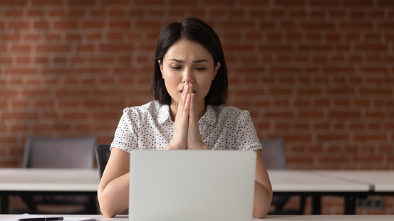 Unhappy Asian businesswoman puts hands in prayer, upset employee asking for help, praying with hope, using laptop, looking at screen, working on difficult task or receive bad news in email