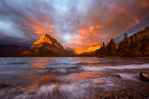 Fire blazes across the sky and a rainbow appears as a late season storm powers its way onto Swiftcurrent Lake raising the water into froth and wave in one of the most beautful places on earth, Glacier National Park located in Montana.