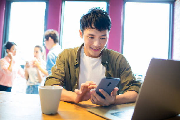man use smartphone and laptop asian young man use 5g smartphone and laptop in the restaurant male chinese student stock pictures, royalty-free photos & images