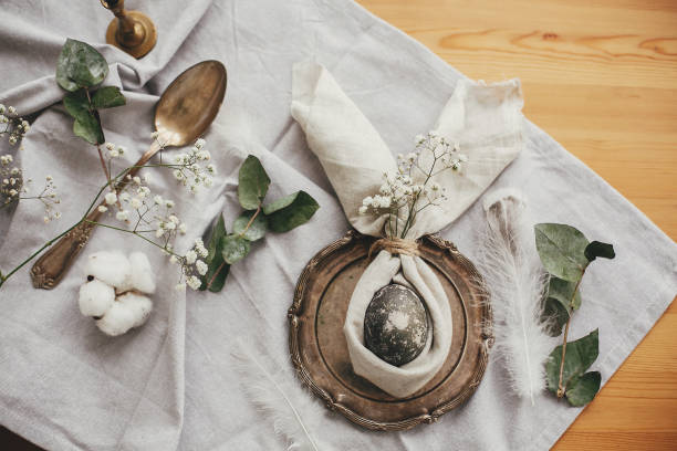 istock Stylish Easter brunch table setting with egg in easter bunny napkin. Modern natural dyed gray egg on napkin with bunny ears, flowers on vintage plate and cutlery. Easter table decorations 1191364633