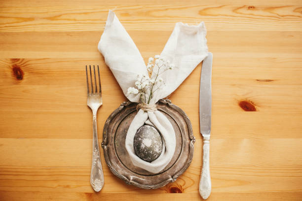 istock Stylish Easter brunch table setting with egg in easter bunny napkin. Modern natural dyed gray egg on napkin with bunny ears, flowers on vintage plate and cutlery. Easter table decorations 1191364704