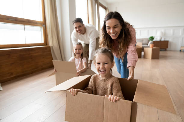 Happy parents playing with little kids riding in box Happy parents playing with cute small kids daughters laughing on moving day, family tenants renters homeowners and children girls having fun riding in box in living room relocating new home concept ecstatic photos stock pictures, royalty-free photos & images