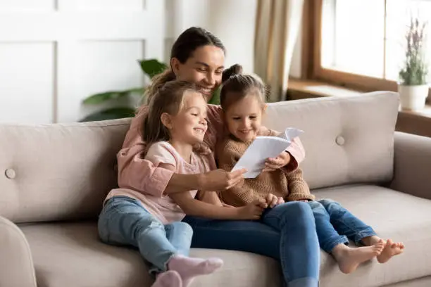 Happy family young mother babysitter hold read book relax embrace cute little children daughters, smiling parent mum tell small kids funny fairy tale story sit on sofa having fun together at home