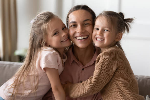 Happy mother and children daughters embracing looking at camera Beautiful female family young smiling single mother and cute little children daughters embracing looking at camera, funny small kids girls hugging happy mum bonding sit on sofa together, portrait teeth bonding stock pictures, royalty-free photos & images