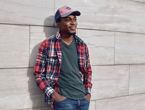 Young smiling african man looking away wearing casuals, red plaid shirt, baseball cap on city street over gray brick wall background