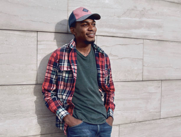 Young smiling african man looking away wearing casuals, red plaid shirt, baseball cap on city street over gray brick wall background stock photo