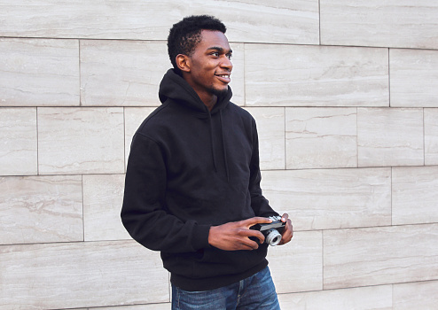 Smiling african man photographer with vintage film camera looking away wearing black hoodie while walking on city street over gray wall background