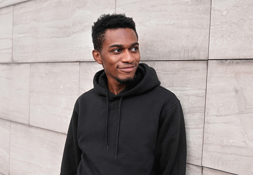 Portrait smiling young african man wearing black hoodie on city street over gray wall background