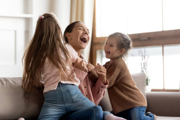 Small kids daughters tickling playing with happy mum on sofa Cheerful positive family cute small funny kids children daughters enjoying tickling playing lifestyle game with young happy mum embracing laughing having fun relaxing sitting on sofa at home together happy sibling day stock pictures, royalty-free photos & images