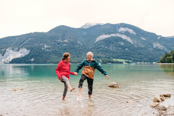 um pensionista sênior pares caminhantes que estão descalços no lago na natureza. - senior couple senior adult action retirement - fotografias e filmes do acervo