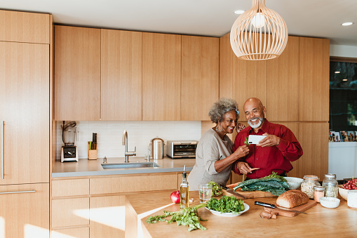 Smiling senior couple watching video while preparing vegan meal in kitchen. Retired man and woman are standing with vegetable at kitchen island. They are spending leisure time at home.