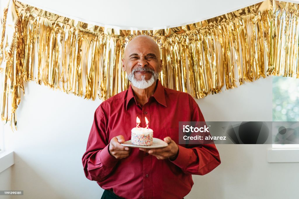 Man holding birthday cake with burning candles Portrait of smiling senior man holding birthday cake with burning candles. Retired male is standing with dessert against decoration. He is making face at home. Birthday Stock Photo