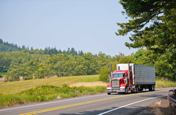 Big rig red classic powerful semi truck with vertical pipes transporting cargo in refrigerator semi trailer running on the road with meadow and trees on the hill
