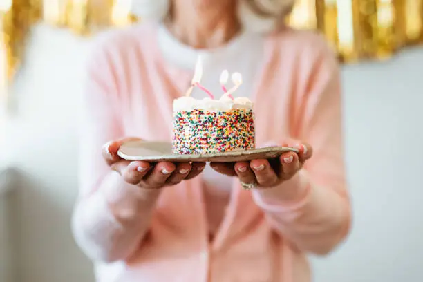 Midsection of retired woman holding cake with burning birthday candles. Senior female is standing with dessert in plate. She is celebrating at home.